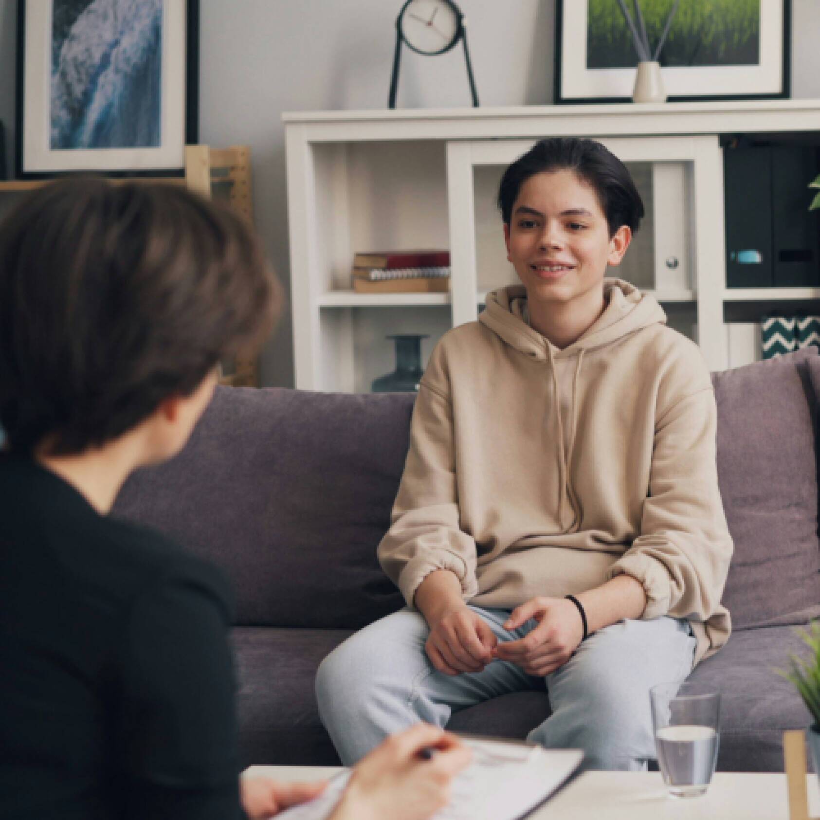 A psychologist has her back to the camera in the foreground, talking to an adolescent in a tan hoodie. Photo from UnSplash by Vitaly Gariev.